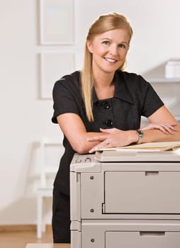 A businesswoman is standing in an office near the copy machine.  She is smiling at the camera.  Vertically framed shot.
