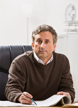 Attractive male sitting at desk with pen and paper, looking at camera. Vertical