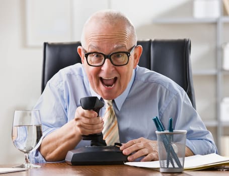 An elderly businessman is seated at a desk in his office and is joking around.  He is looking at a camera.  Horizontally framed shot.