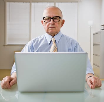 An elderly businessman is working on a laptop in an office.  He is looking at the camera.  Square framed shot.