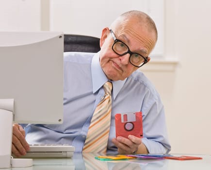 An elderly businessman is seated in an office holding a floppy disk.  He is looking at the camera.  Horizontally framed shot.