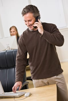 Attractive business man standing over keyboard while chatting on the phone. Woman peering in back. Looking at the computer screen. Vertical.