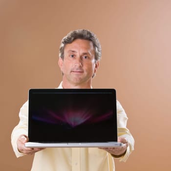 A man is holding a laptop out in front of him and looking at the camera.  Square framed shot.