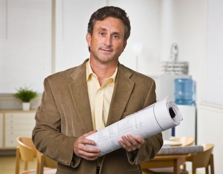 A businessman is standing in an office, holding blueprints, and looking at the camera.  Horizontally framed shot.