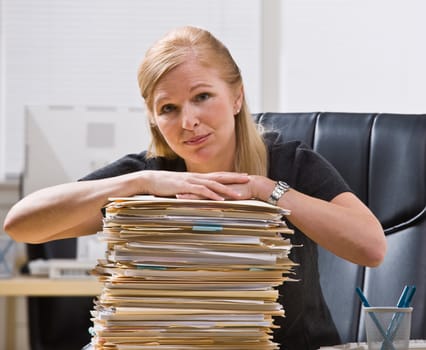 A businesswoman is seated at a desk with a large stack of paperwork in front of her.  She is looking at the camera.  Horizontally framed shot.