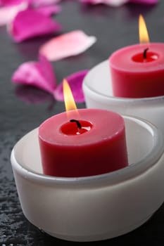 A close-up of a health and beauty spa decoration with two red colored candles in white opaque bowls surrounded by pink flower petals on a black stone surface.