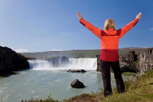 A young woman with her arms up in celebration of achievement by a waterfall. Shot on location at Godafoss waterfall in Iceland.