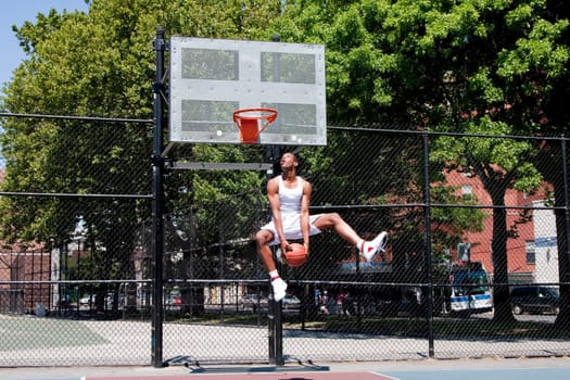 Sporty handsome African-American man dressed in white jumping in the air reaching for the basket while playing basketball in an outdoor court on a summer day