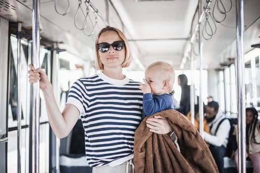 Mother carries her child while standing and holding on to bar holder on bus. Mom holding infant baby boy in her arms while riding in public transportation. Cute toddler traveling with mother