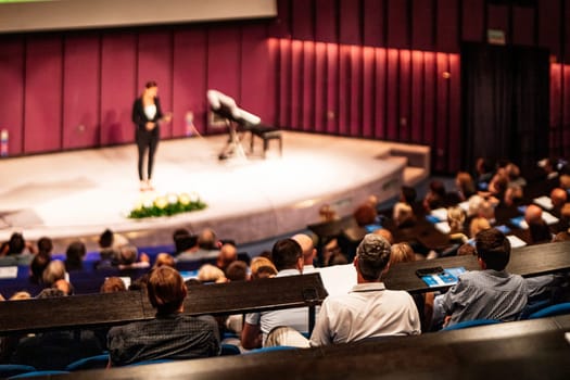Business and entrepreneurship symposium. Female speaker giving a talk at business meeting. Audience in conference hall. Rear view of unrecognized participant in audience.