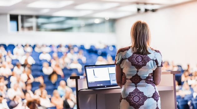 Female speaker giving a talk on corporate business conference. Unrecognizable people in audience at conference hall. Business and Entrepreneurship event.
