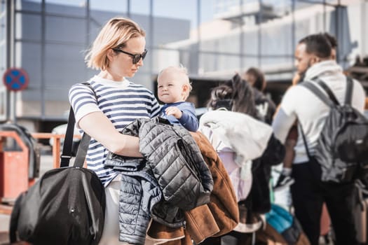 Motherat travelling with his infant baby boy child. Mom holding travel bag and her infant baby boy child while queuing for bus in front of airport terminal station.