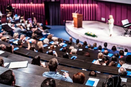 Business and entrepreneurship symposium. Female speaker giving a talk at business meeting. Audience in conference hall. Rear view of unrecognized participant in audience.