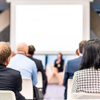 Business and entrepreneurship symposium. Female speaker giving a talk at business meeting. Audience in conference hall. Rear view of unrecognized participant in audience.