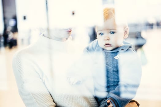 Thoughtful young mother looking trough window holding his infant baby boy child while waiting to board an airplane at airport terminal departure gates. Travel with baby concept