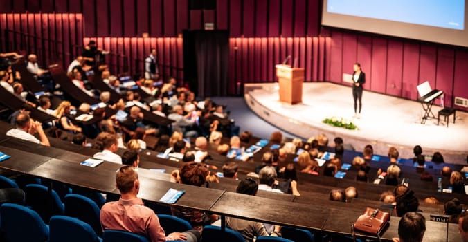 Business and entrepreneurship symposium. Female speaker giving a talk at business meeting. Audience in conference hall. Rear view of unrecognized participant in audience.
