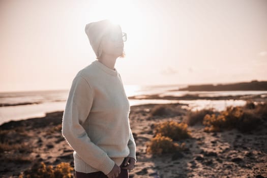Portrait of young stylish woman wearing wool sweater, wool cap and sunglasses on long sandy beach in spring
