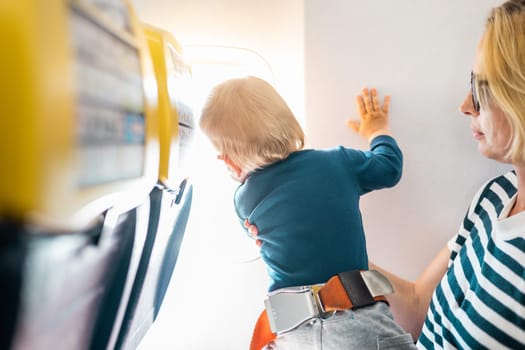 Mom and child flying by plane. Mother holding and playing with her infant baby boy child in her lap during economy comercial flight. Concept photo of air travel with baby. Real people