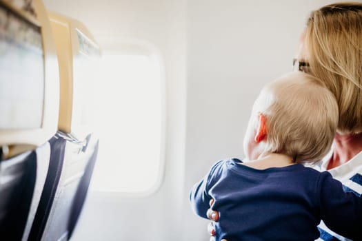 Mom and child flying by plane. Mother holding and playing with her infant baby boy child in her lap during economy comercial flight. Concept photo of air travel with baby. Real people