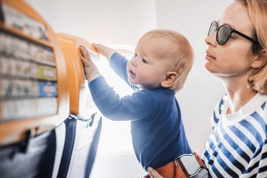 Mom and child flying by plane. Mother holding and playing with her infant baby boy child in her lap during economy comercial flight. Concept photo of air travel with baby. Real people