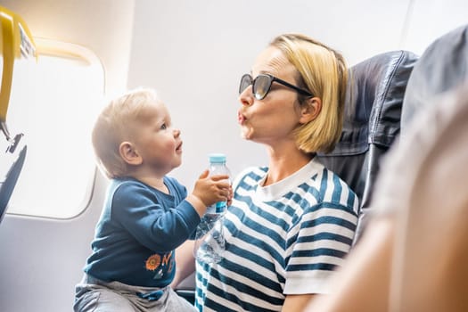 Mom and child flying by plane. Mother holding and playing with her infant baby boy child in her lap during economy comercial flight. Concept photo of air travel with baby. Real people