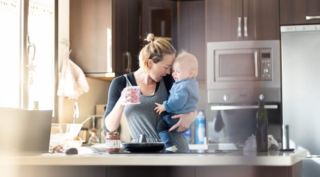 Happy mother and little infant baby boy together making pancakes for breakfast in domestic kitchen. Family, lifestyle, domestic life, food, healthy eating and people concept