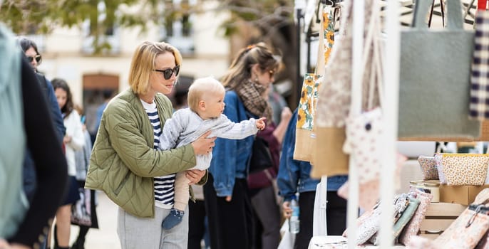 Mother walking carrying his infant baby boy child in crowd of people wisiting sunday flea market in Malaga, Spain.