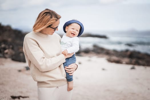 Mother enjoying winter vacations holding and playing with his infant baby boy son on volcanic sandy beach on Lanzarote island, Spain. Family travel and vacations concept.