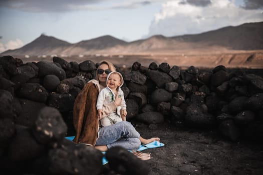 Mother enjoying winter vacations playing with his infant baby boy son on black sandy volcanic beach of Janubio on Lanzarote island, Spain on windy overcast day. Family travel vacations concept