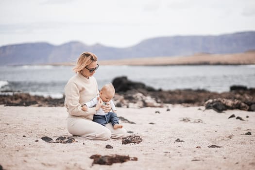 Mother enjoying winter beach vacations playing with his infant baby boy son on wild volcanic sandy beach on Lanzarote island, Canary Islands, Spain. Family travel and vacations concept.