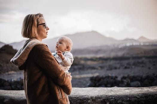 Mother enjoying winter vacations playing with his infant baby boy son on black sandy volcanic beach of Janubio on Lanzarote island, Spain on windy overcast day. Family travel vacations concept
