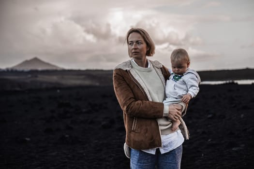 Mother enjoying winter vacations playing with his infant baby boy son on black sandy volcanic beach of Janubio on Lanzarote island, Spain on windy overcast day. Family travel vacations concept