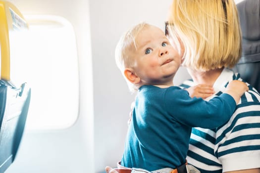 Mom and child flying by plane. Mother holding and playing with her infant baby boy child in her lap during economy comercial flight. Concept photo of air travel with baby. Real people