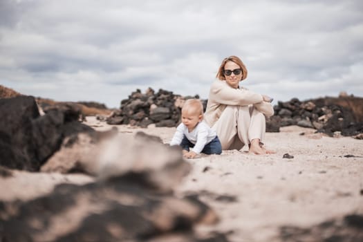 Mother enjoying winter beach vacations playing with his infant baby boy son on wild volcanic sandy beach on Lanzarote island, Canary Islands, Spain. Family travel and vacations concept.