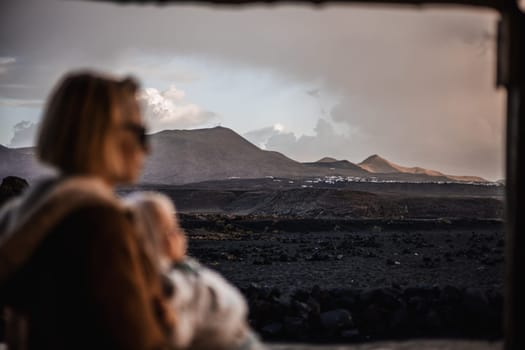 Mother enjoying winter vacations playing with his infant baby boy son on black sandy volcanic beach of Janubio on Lanzarote island, Spain on windy overcast day. Focus on landscape in background