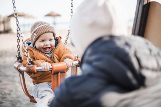 Mother pushing her cheerful infant baby boy child on a swing on sandy beach playground outdoors on nice sunny cold winter day in Malaga, Spain