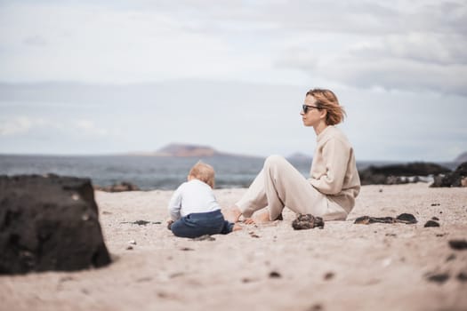 Mother enjoying winter beach vacations playing with his infant baby boy son on wild volcanic sandy beach on Lanzarote island, Canary Islands, Spain. Family travel and vacations concept.