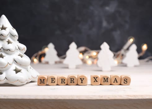 Small wooden blocks with Merry Christmas and white wooden Christmas tree, blurred lights in the background