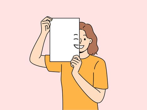 Smiling woman holding piece of paper with emoji in front of face and looking at screen. Positive girl with partially painted smile is sharing good mood and positive emotions with you.
