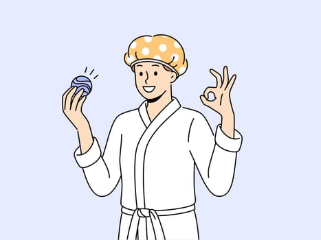 Man with bath bomb is dressed in bathrobe and bandage to protect hair from moisture. Young guy shows OK gesture recommending use of aromatic bath bomb while performing hygiene procedures.