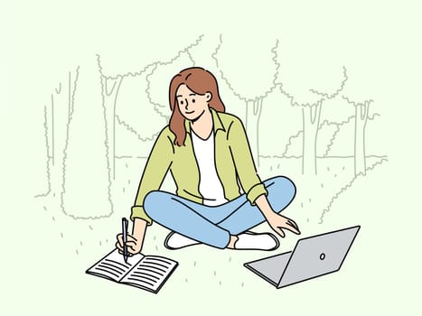 Woman student is sitting on lawn making notes in notebook and using laptop enjoying working in park. Happy girl student watching educational video and writing notes located in park