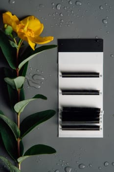 View from above of professional palette of black artificial eyelashes for lash extension procedure lying on gray table surrounded by drops of water, near yellow flower. Vertical.