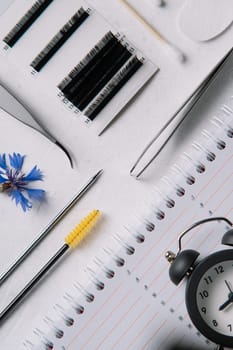 View from above of various tools and materials for eyelash extension procedure, yellow brush, tweezers and palette of cilia, lying on white table near blue cornflower and diary with clocks. Vertical.