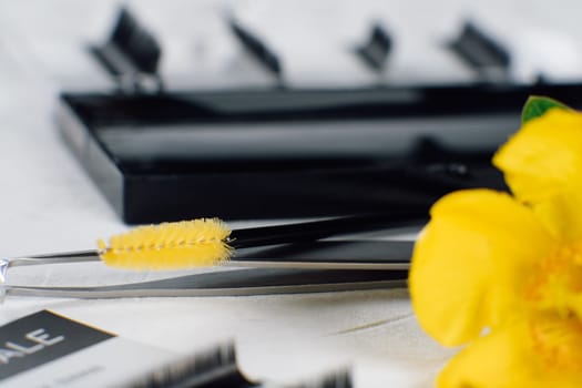 Bright flower, left on foreground, slightly covering disposable eyelash extension brush with yellow delicate bristles lying on isolated tweezers on white table full of tools for procedure.