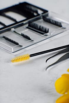 Professional materials and instruments for lash extension procedure, palette of black artificial cilia, disposable brush with yellow bristles and isolated tweezers, lying on white table. Vertical.