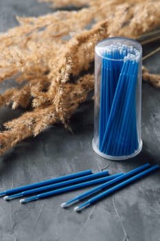 Professional microbrushes with blue stick lying in container and left on gray table near branches of beige pampas grass. Tool for eyelash extension procedure using as applicator. Vertical.