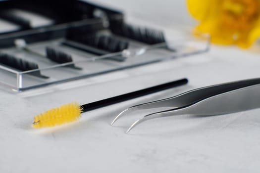 Pair of various tools for separating lashes throughout eyelash extension procedure, yellow disposable brush and isolated tweezers, lying on white table in front of palette of black artificial cilia.