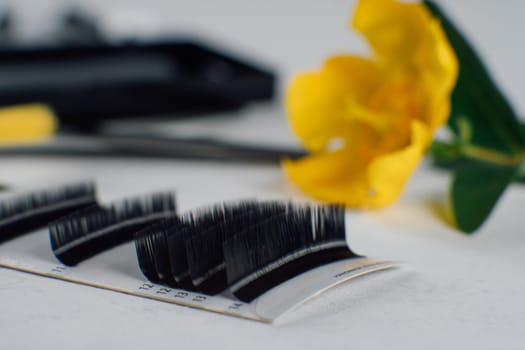Professional palette of black artificial eyelashes lying on white table in front of yellow blossom. Fluffy material applying on eyelids and creating fashionable look in process of cilia extension.