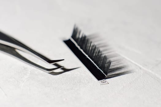 Tweezers with sharp tip, using for isolating small lashes, lying near palette of black artificial eyelashes on white table with cast shandows of tools. Instruments for cilia extension procedure.
