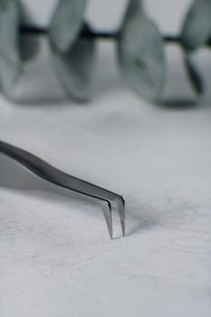 Close up of sharp tip of tweezers for separating and sticking false eyelashes throughout lash extension procedure, lying on white table in front of stem of plant. Vertical.